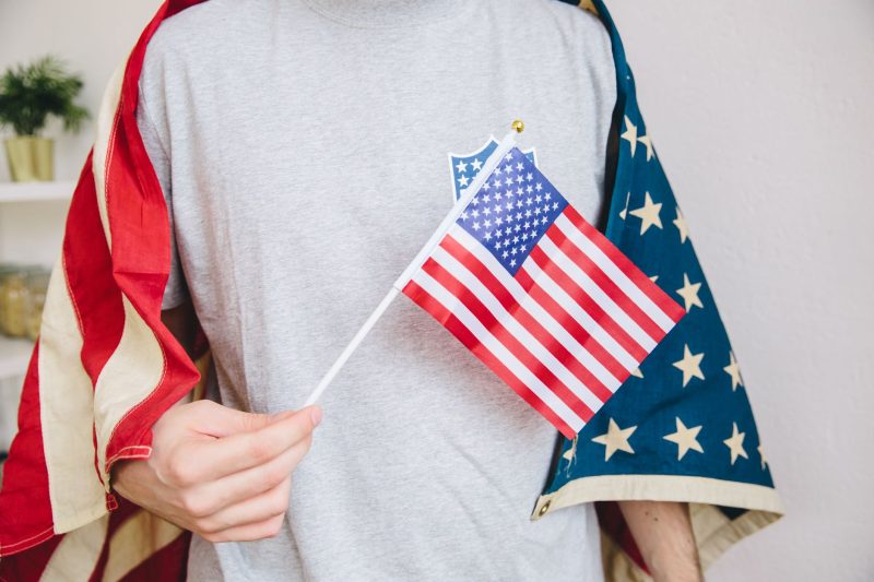 Person indoors holding a small American flag, surrounded by a larger US flag draped around them.